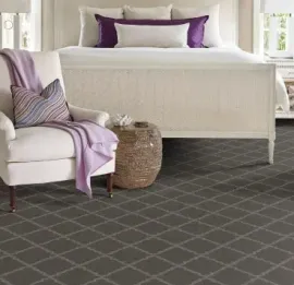 A bedroom features a white upholstered chair with a purple throw, a woven side table, and a patterned dark gray carpet.