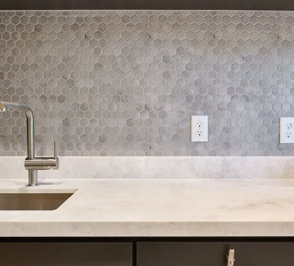 Modern kitchen sink with a chrome faucet, light stone countertop, and a gray hexagonal tile backsplash.