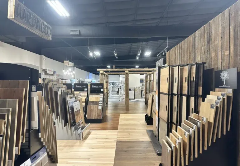 A showroom interior displaying numerous wood flooring samples and planks on racks, with a central aisle.