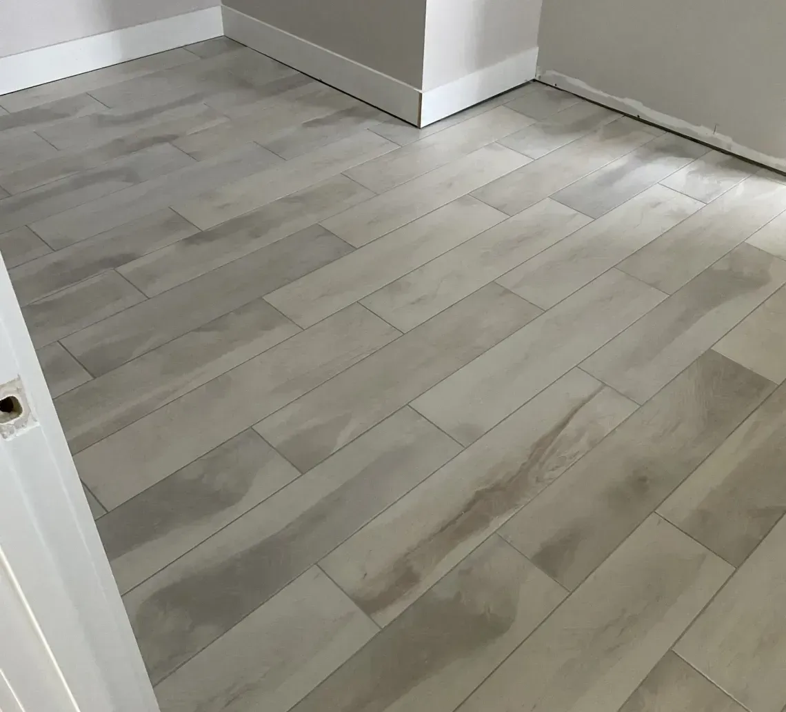 Light-colored wood-look floor tiles installed in a room with white baseboards and grey walls.
