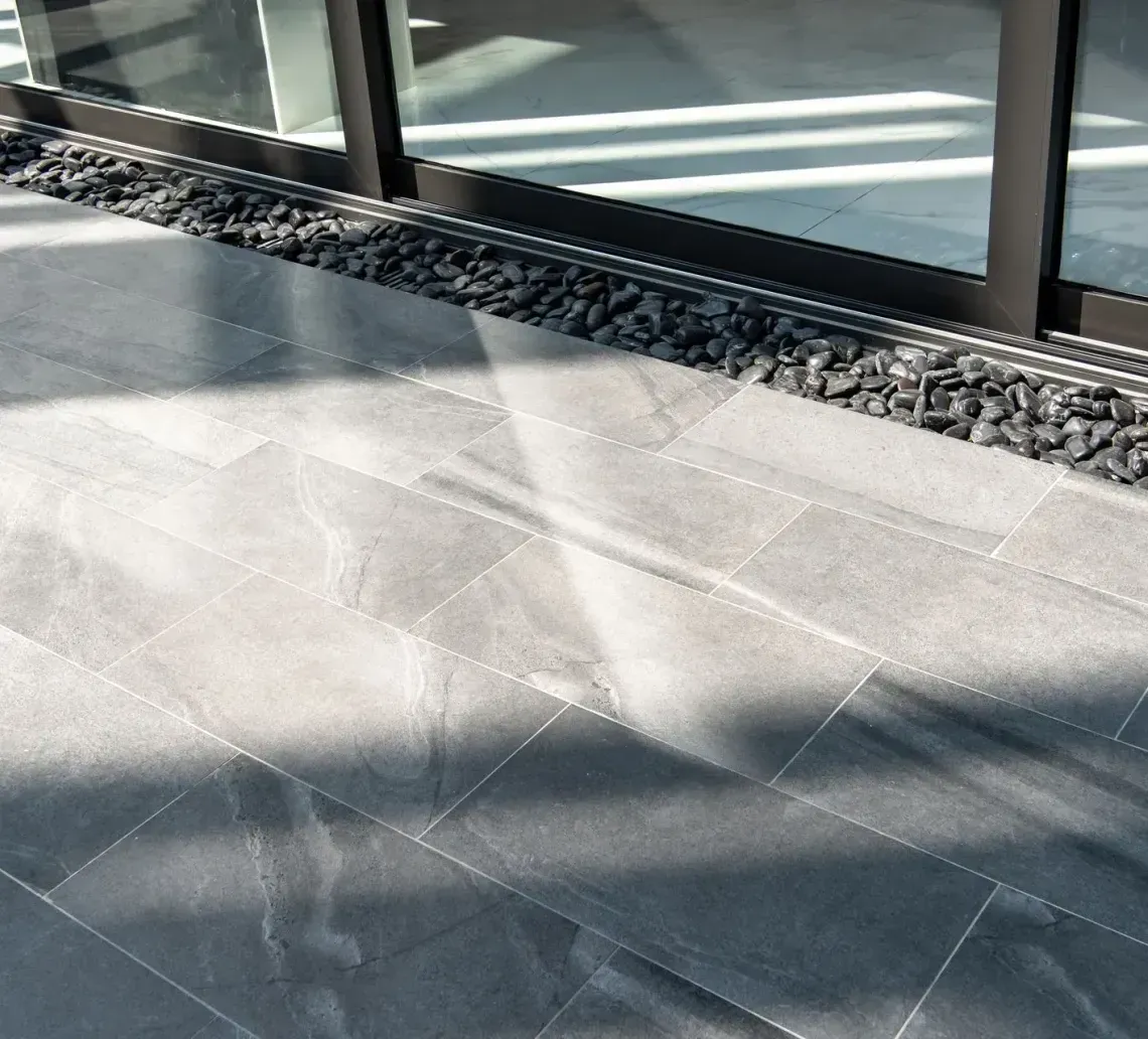 A close-up of grey rectangular patio pavers adjacent to a strip of black landscape rocks and a dark-framed glass door.
