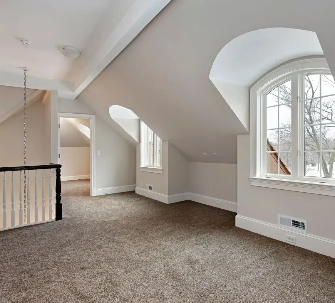 An attic room with neutral walls, tan carpet, two dormer windows, and a staircase with a black railing.