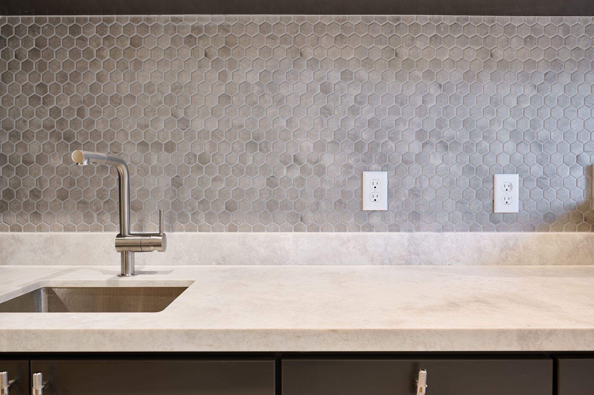 Modern kitchen sink with a chrome faucet, light stone countertop, and a gray hexagonal tile backsplash.