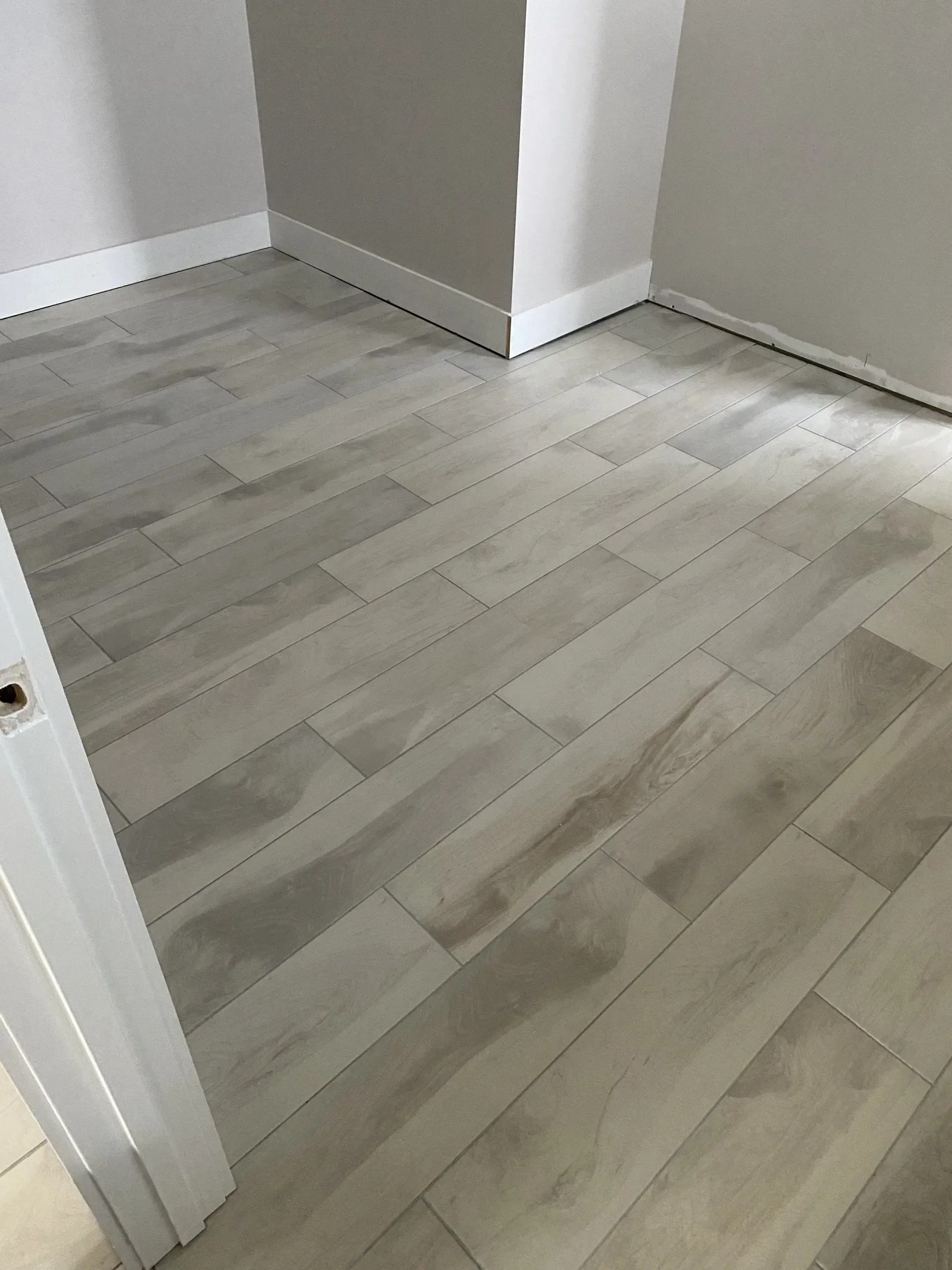 Light-colored wood-look floor tiles installed in a room with white baseboards and grey walls.
