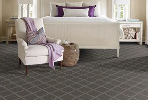 A bedroom with a light-colored armchair and throw blanket, a bed with purple pillows, and dark grey plaid-patterned carpet.