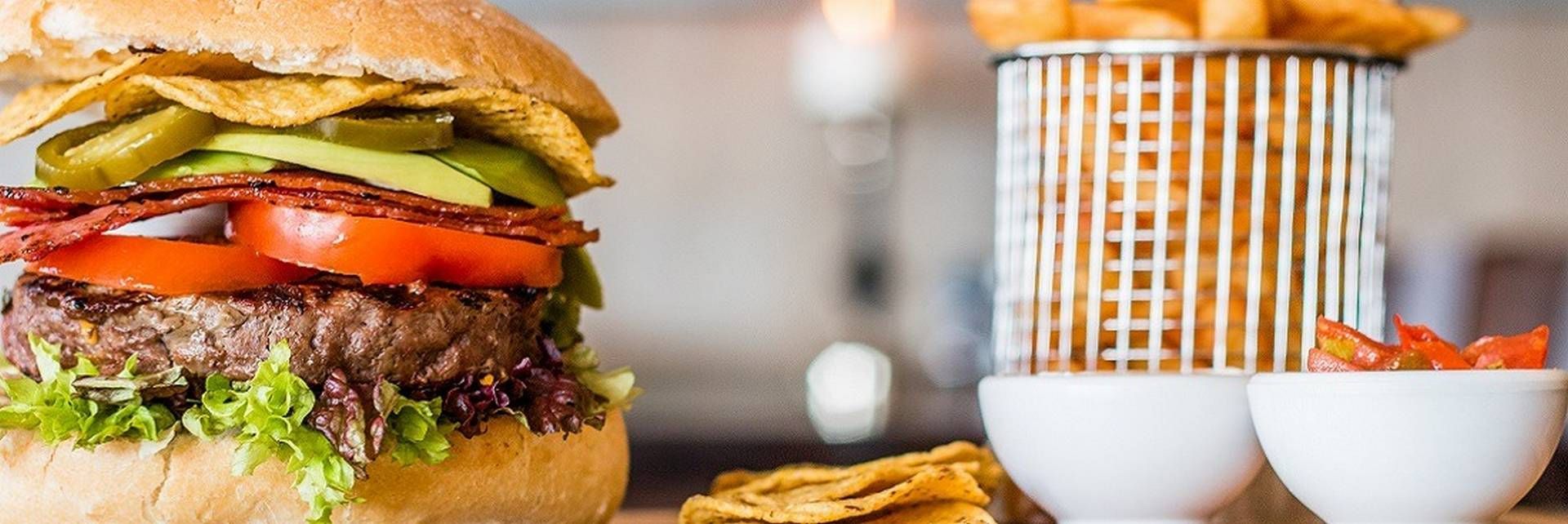 Een luxe burger met sla, tomaat en nacho's, geserveerd met een metalen mandje friet en bijgerechten op tafel.