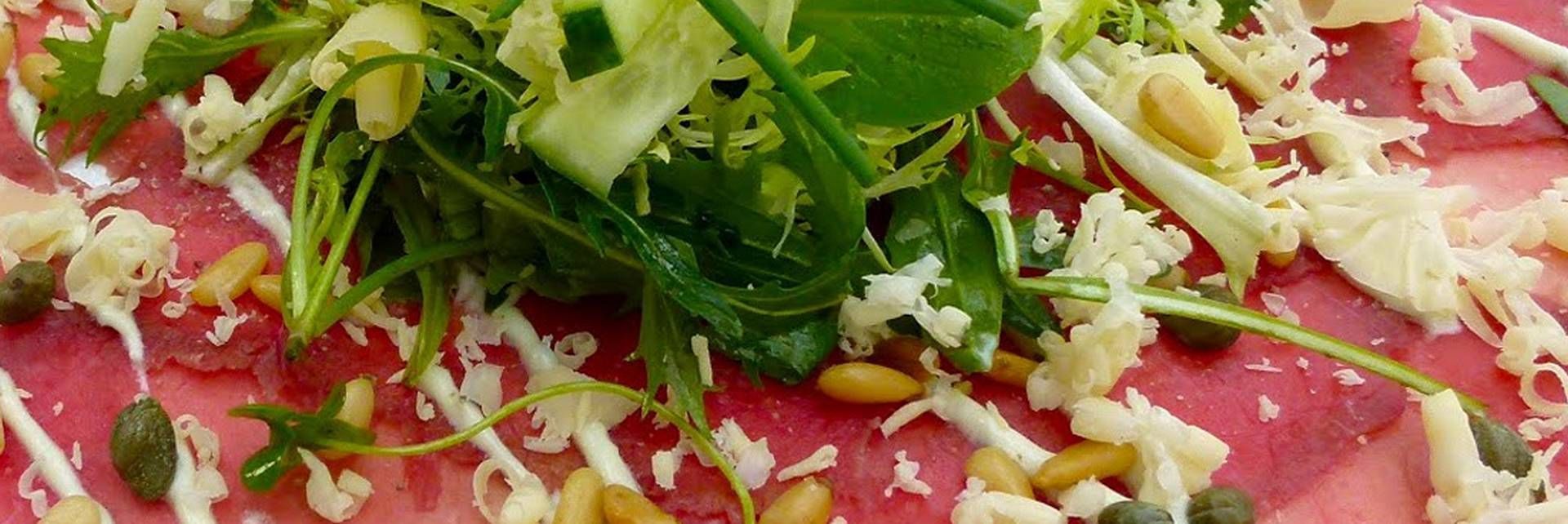 Dun gesneden rundercarpaccio, gegarneerd met rucola, pijnboompitten, kappertjes, geraspte kaas en een lichte scheut saus.