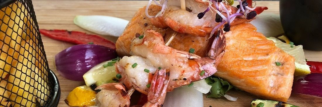 Gegrilde zalm en garnalen geserveerd op een houten plank met kleurrijke groenten en een portie friet in een draadmandje.