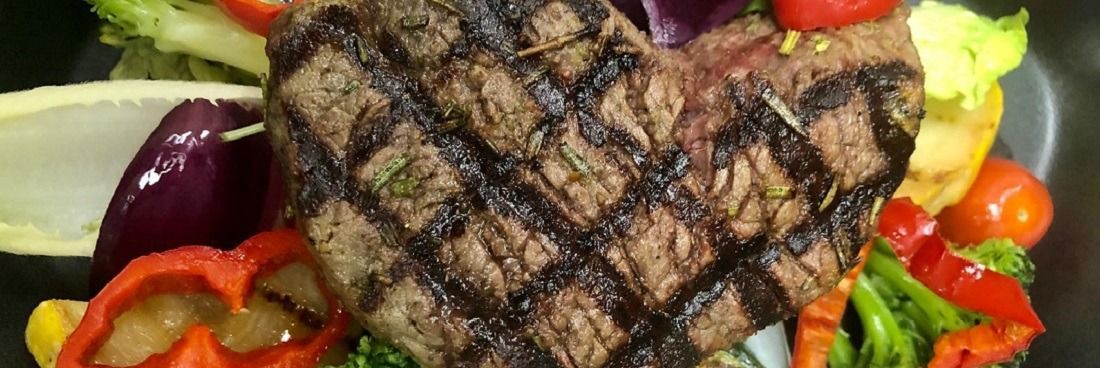 Gegrilde hartvormige biefstuk gegarneerd met geroosterde groenten, waaronder paprika, broccoli en een schijfje citroen.