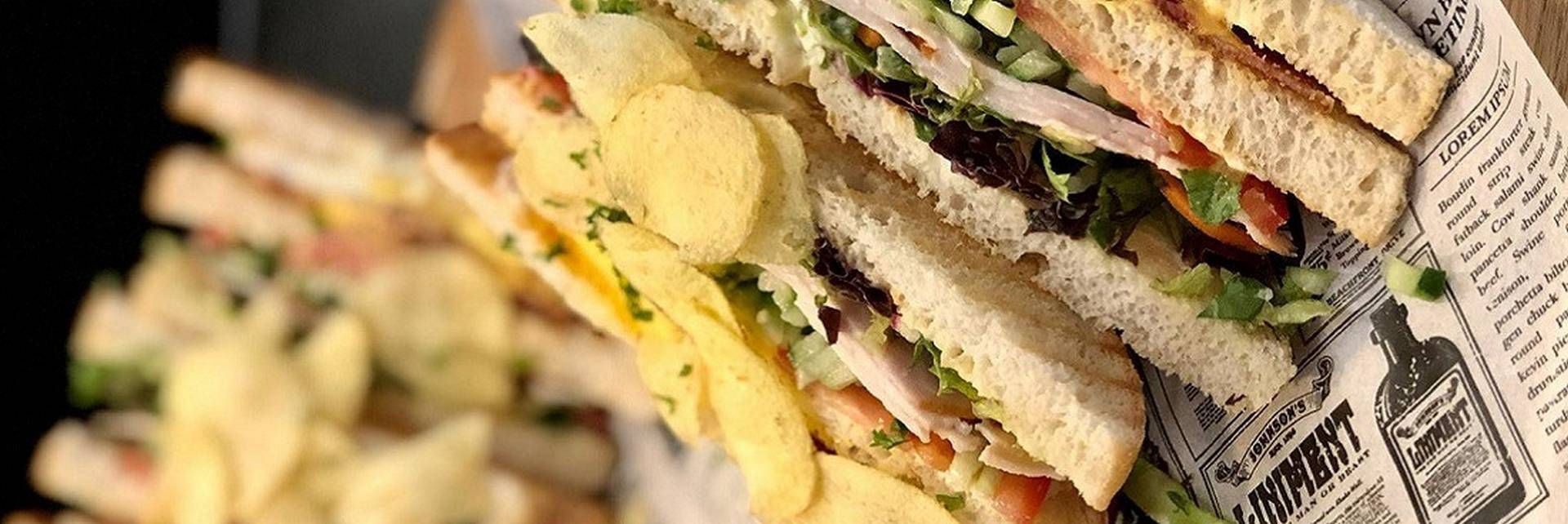 Een close-up van een gestapelde sandwich, geserveerd met chips op perkamentpapier met een vintage tekst.