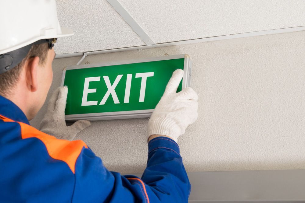 A Man is Installing an Exit Sign on the Wall — Mick Sawtell Electrical Pty Ltd in South Lismore, NSW