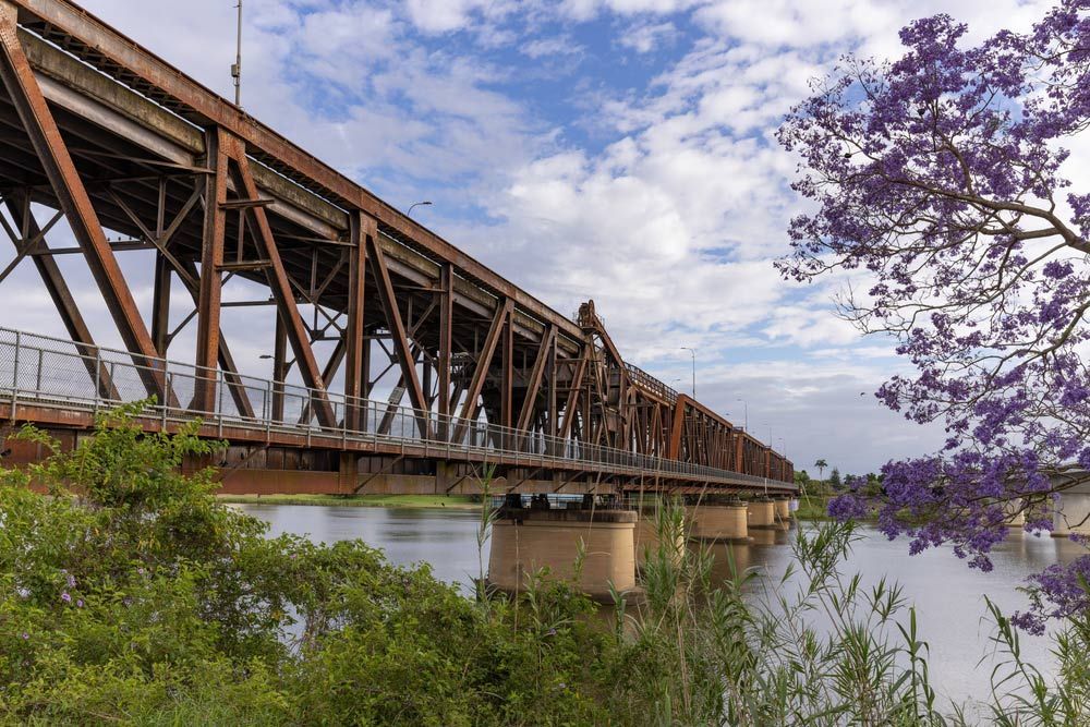 A Bridge Over a River With Purple Flowers in the Foreground — Mick Sawtell Electrical Pty Ltd in Grafton, NSW