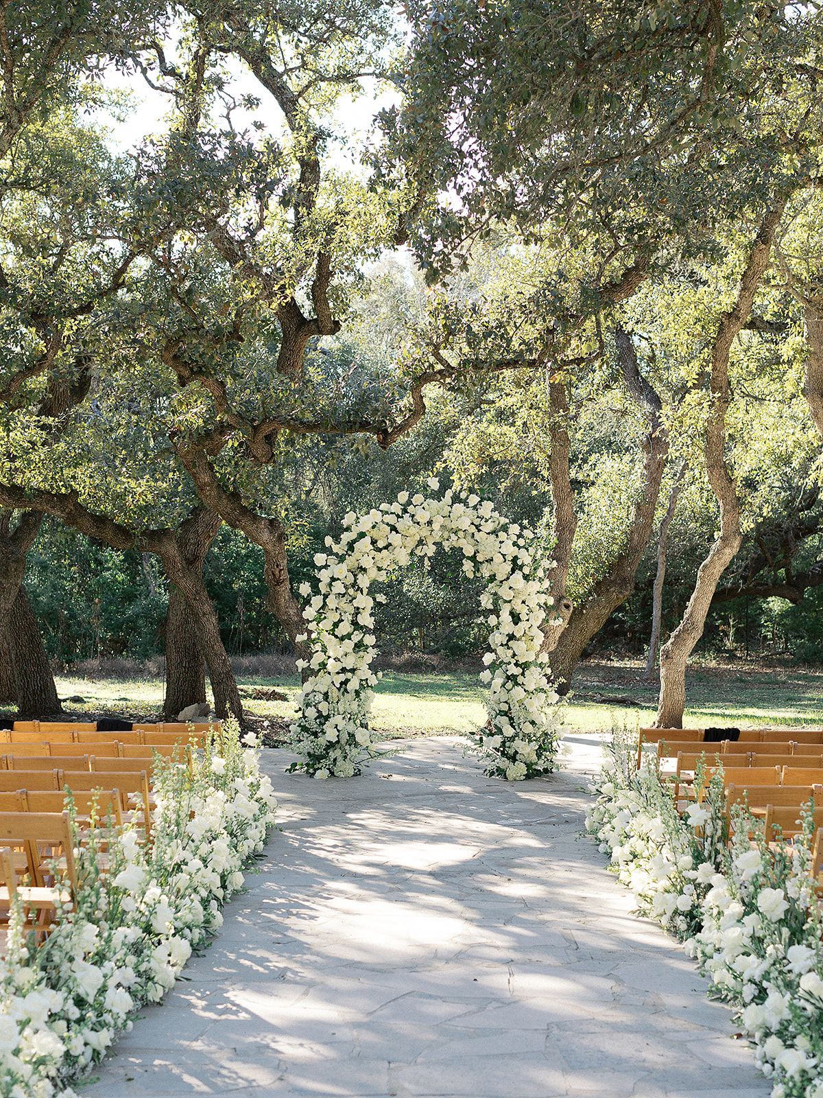 A wedding ceremony is taking place in a park with a floral arch and aisle lined with white flowers.