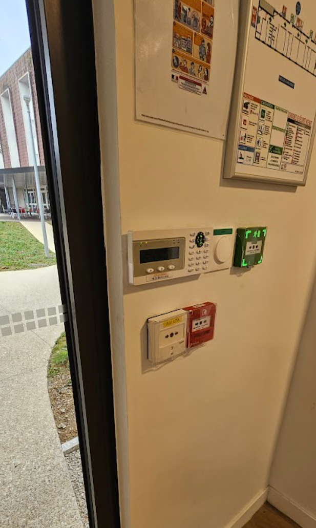 Mur avec alarme incendie, interrupteur, thermostat et panneaux de sortie de secours, près d'une porte.
