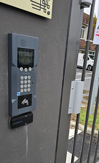Gray intercom system mounted on a gate post with keypad, display, and card reader.