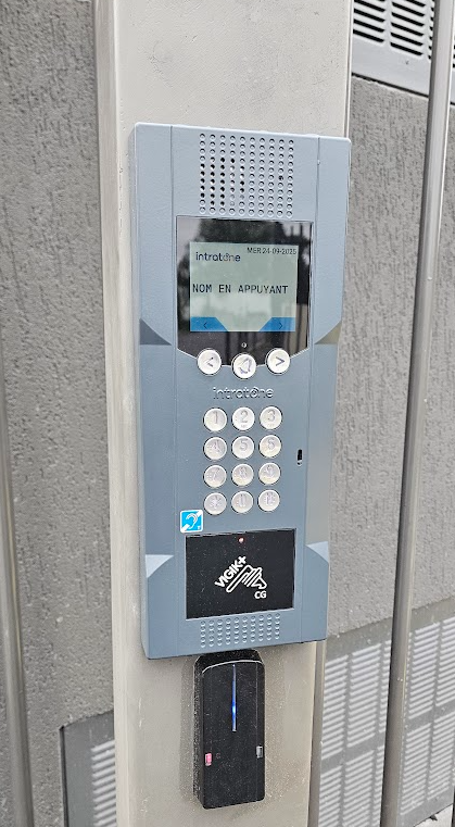 Interphone gris avec clavier, écran et lecteur de cartes, installé sur un pilier en béton.