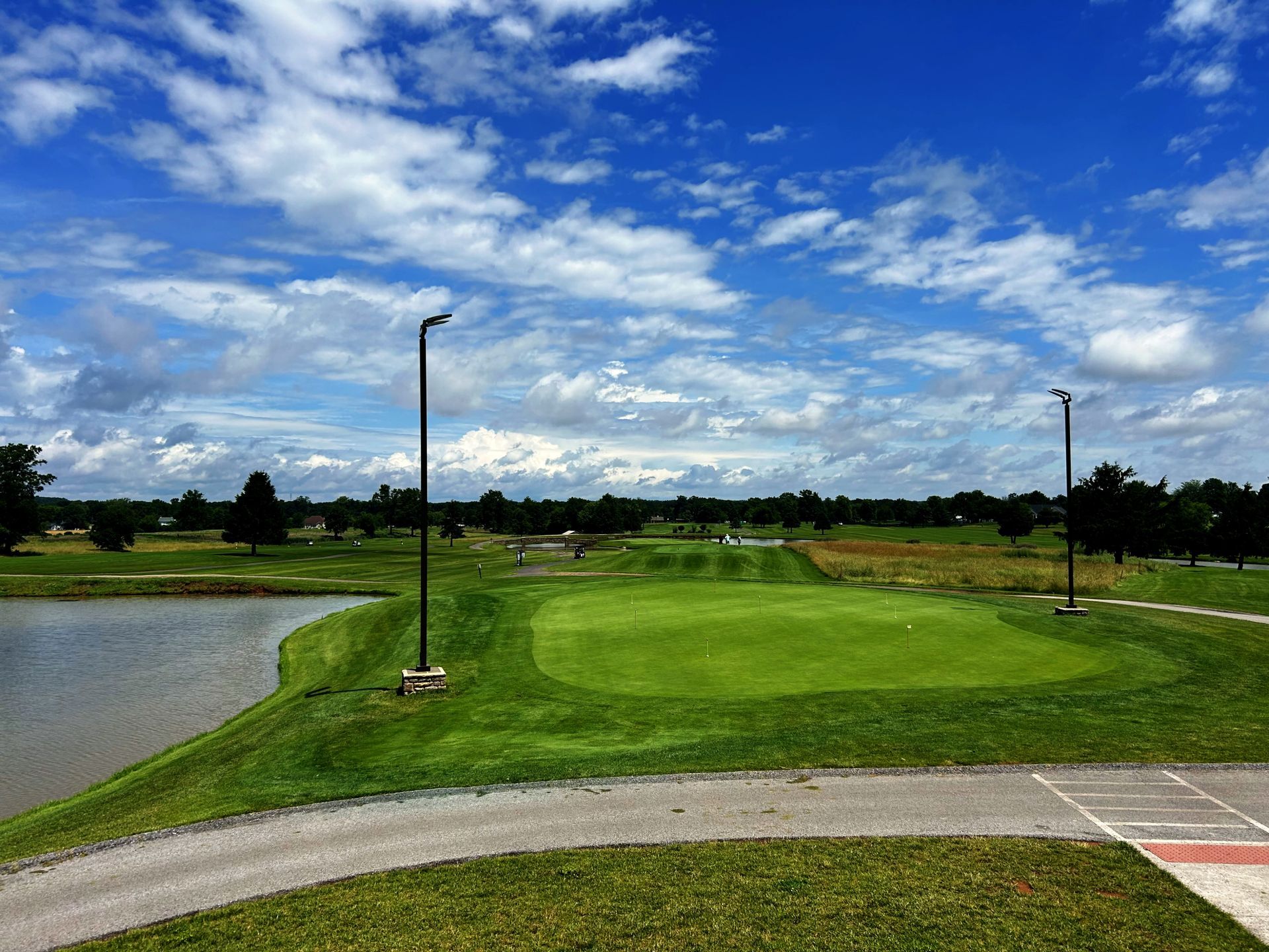 Golf course scene with green grass, lake, blue sky with clouds.