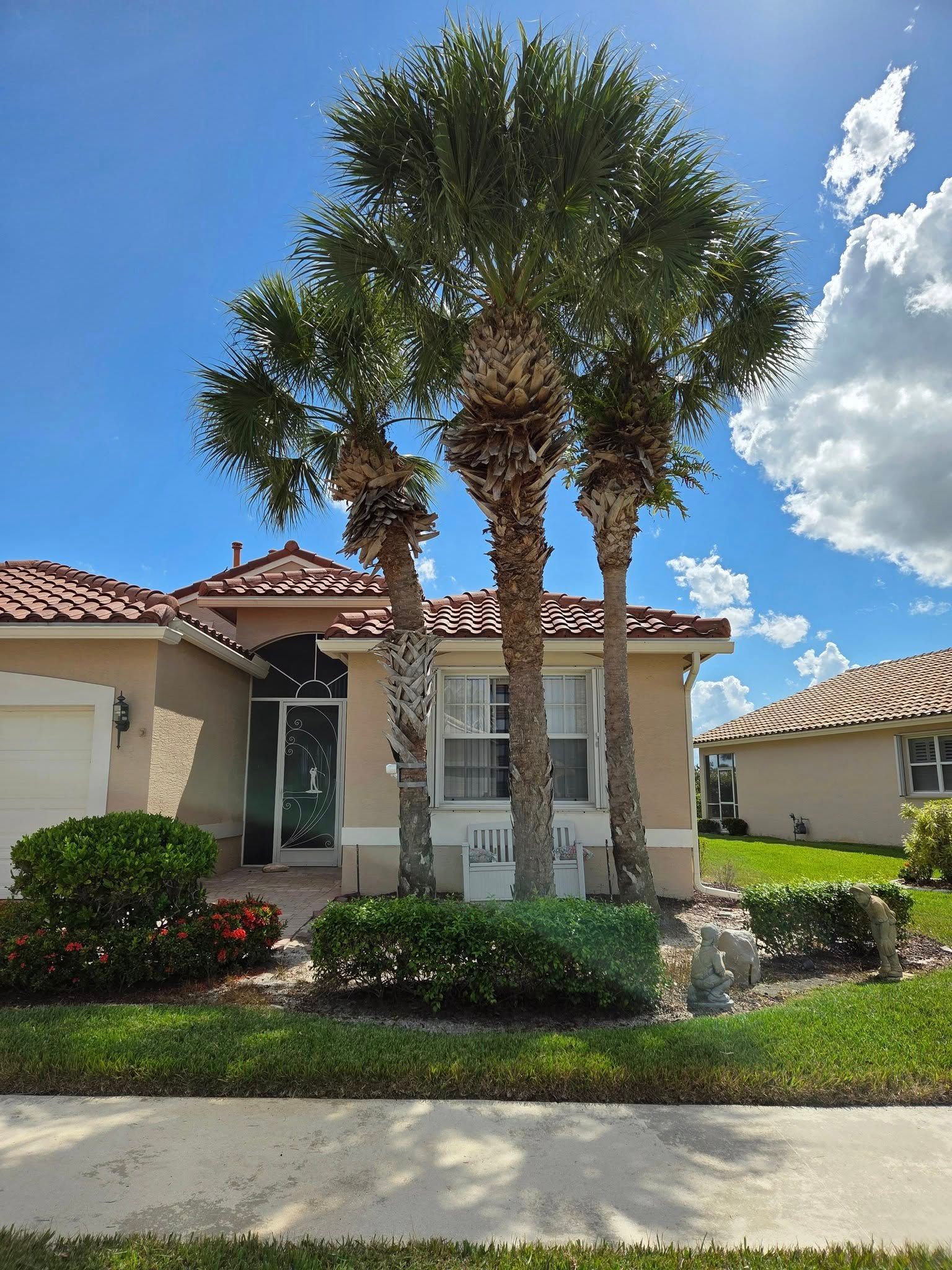 Three palm trees in front of a beige house with a red tile roof, against a blue sky with clouds.