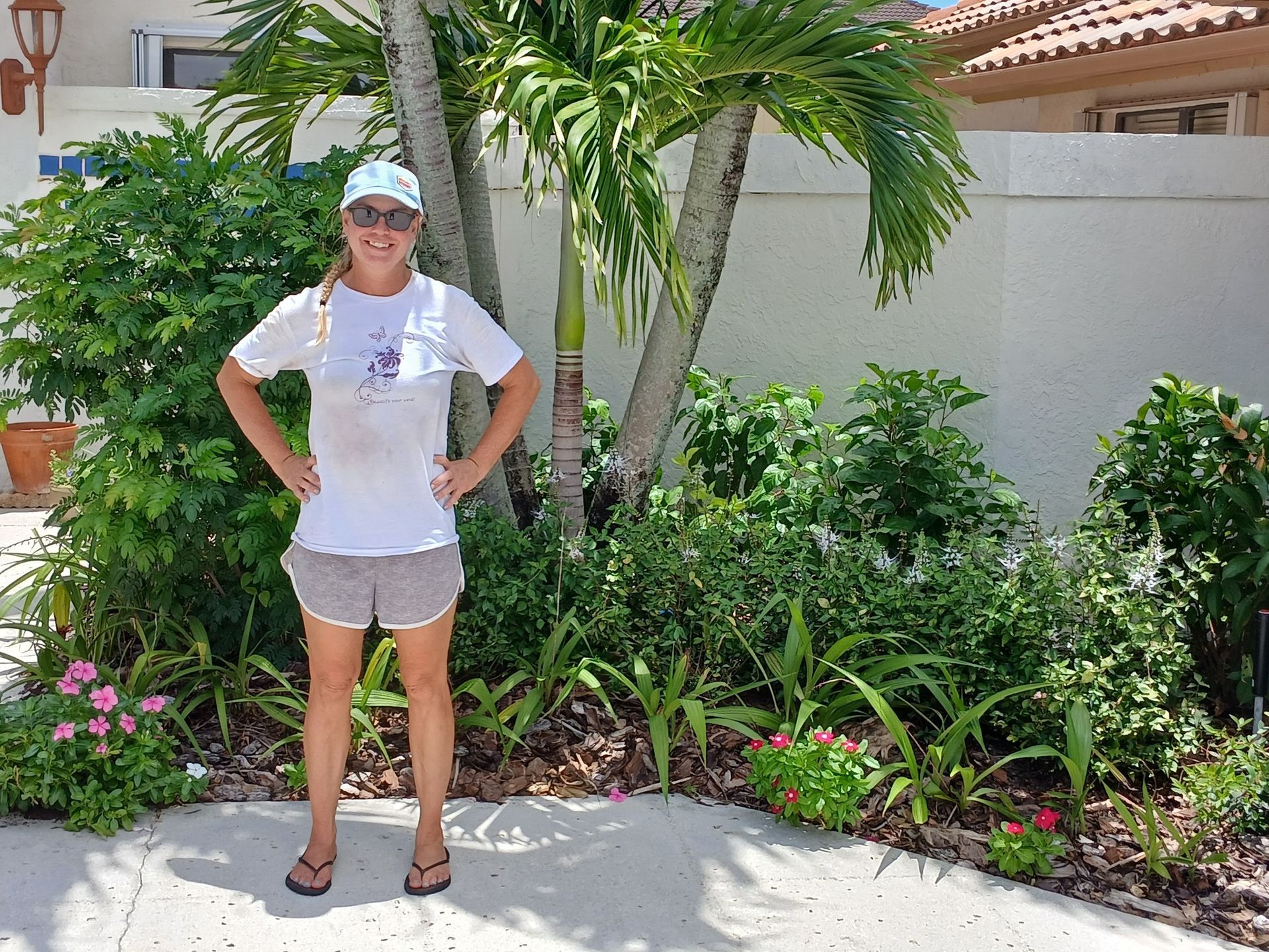 Woman in hat and shorts stands with hands on hips in front of landscaping.