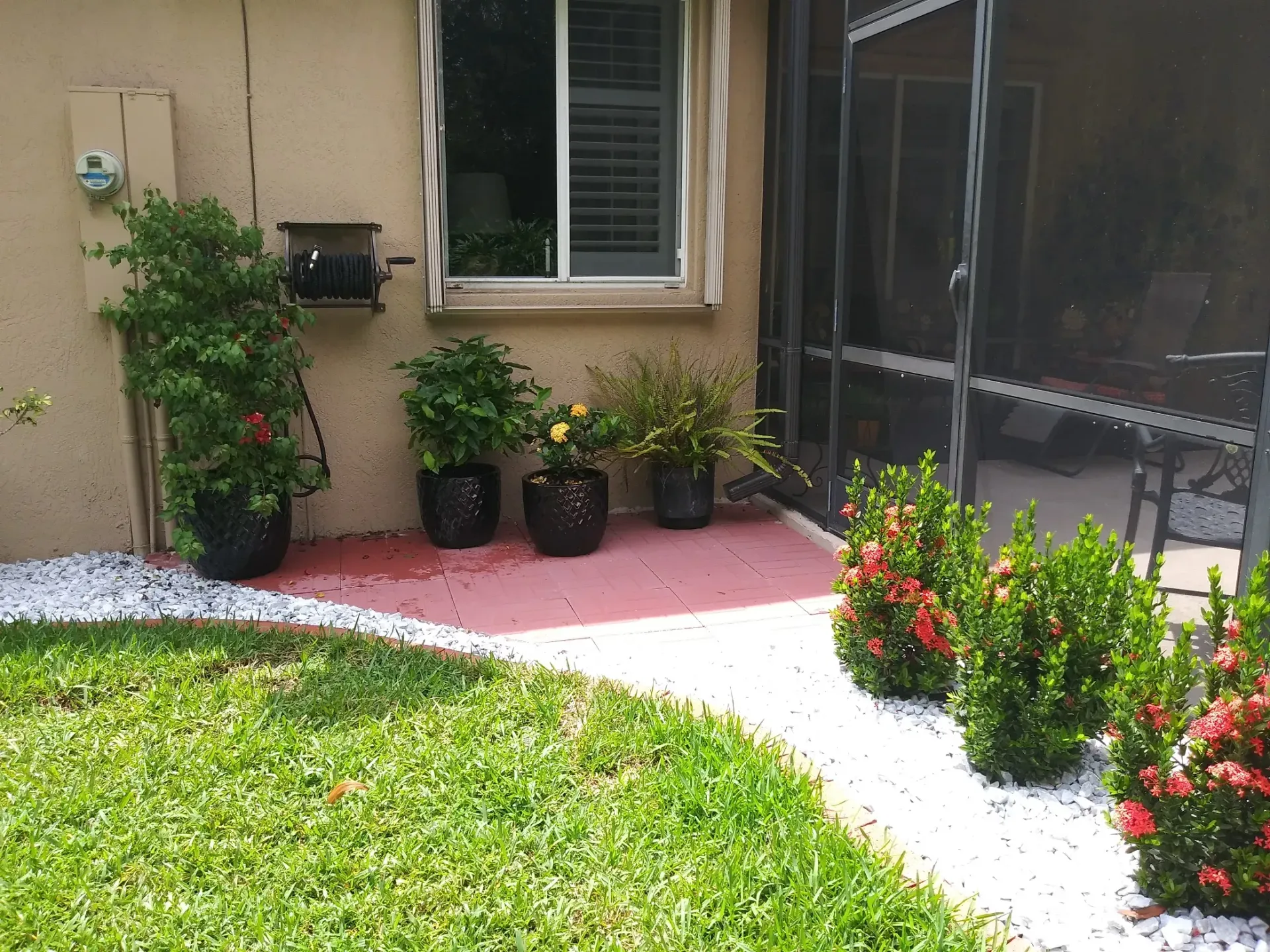 A patio with potted plants, white stones, and green grass. A screened-in porch is to the right.