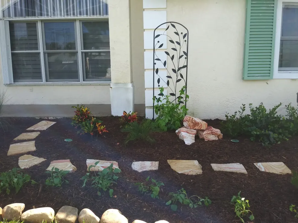 Stone path leads to a house with cream-colored siding and green shutters, a decorative metal panel in the garden.