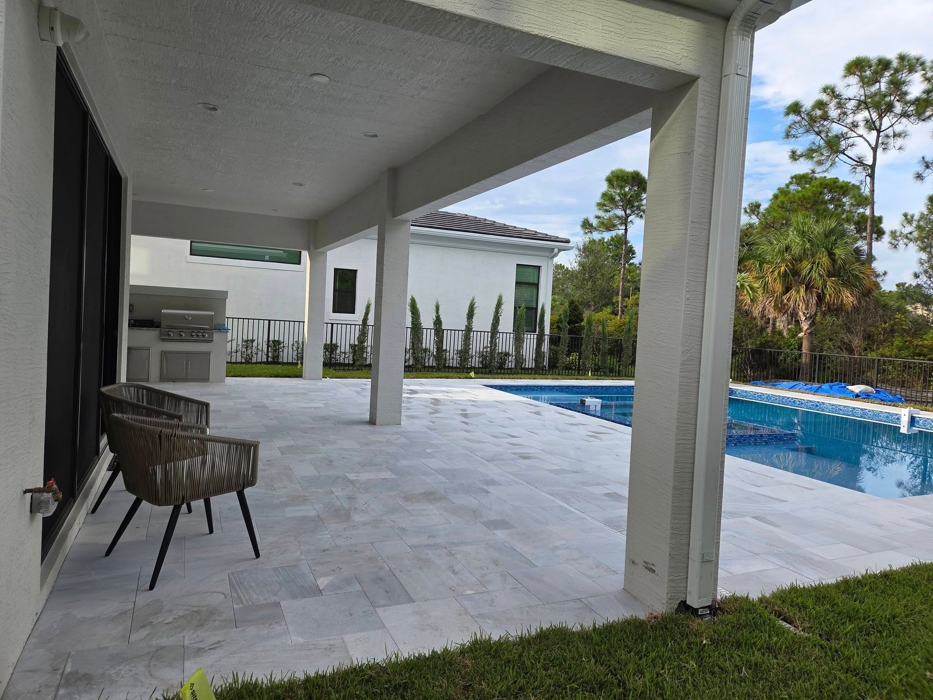 Patio with pool, grill, and seating. White pillars support the roof; lawn and trees in background.