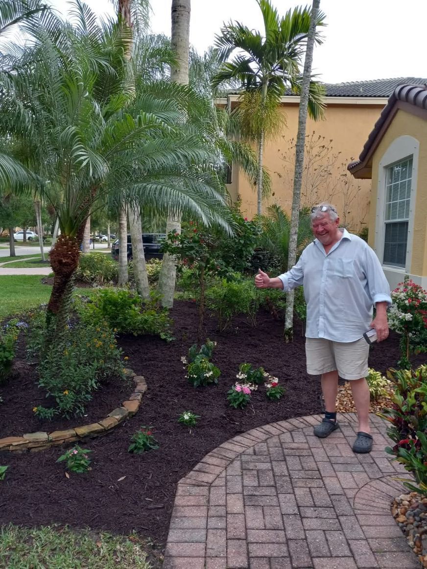 Man with thumb up next to a garden bed with flowers and mulch, in front of a house.