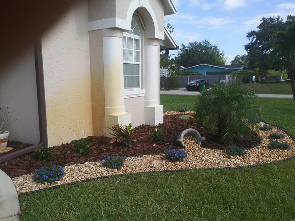 Landscaped yard with flower beds, featuring mulch, river rock, and various plants next to a house with beige stucco.