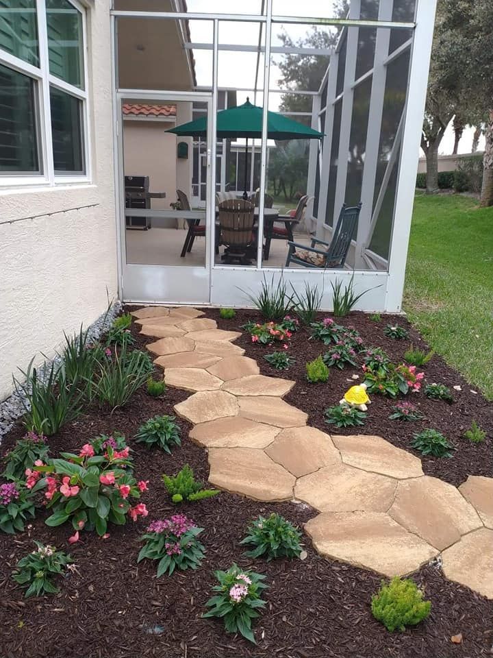 Stone path through a garden with pink and green plants, leading to a screened patio with a green umbrella.