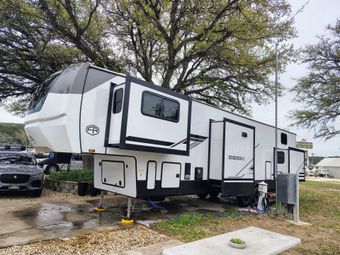 Large white and black fifth-wheel RV parked outdoors on a wet lot under trees
