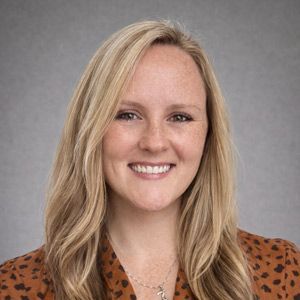 Woman with blonde hair smiling, wearing a patterned orange top, against a gray backdrop.