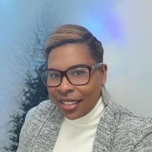 Woman with glasses smiles, wearing a white turtleneck and grey blazer; blurred trees and blue background.