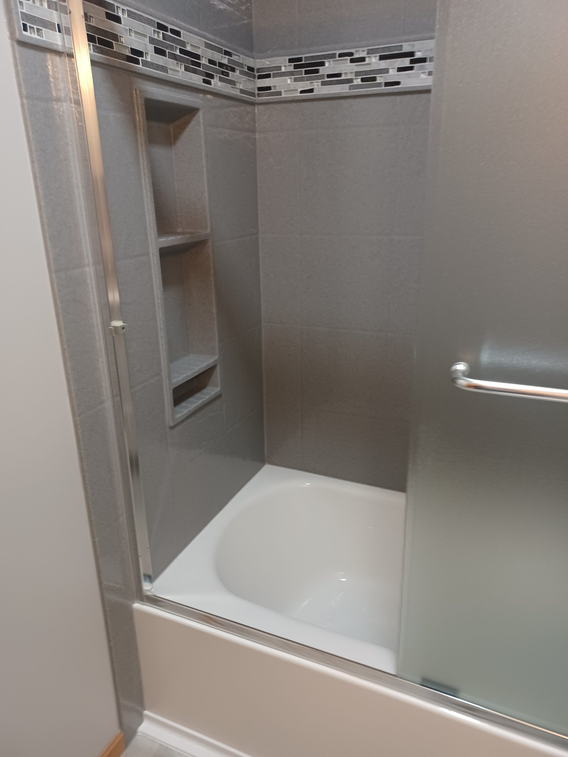 Gray tiled shower stall with glass door, small shelves, and bathtub.