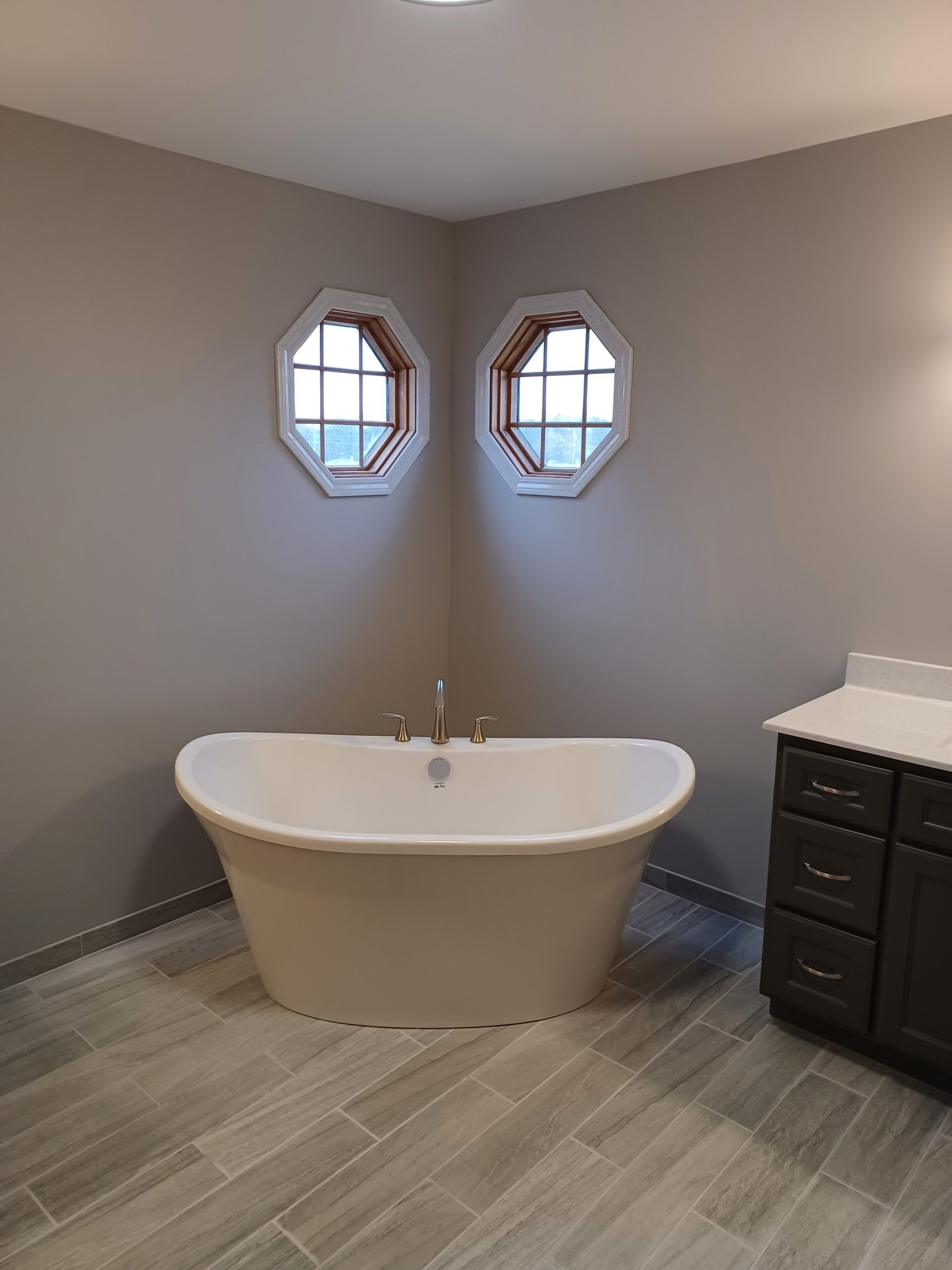 Bathroom with a white freestanding tub between two octagonal windows, gray walls, and dark cabinets.