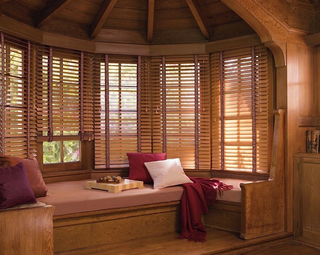 Wooden bay window seat with blinds, pillows, and a blanket. Warm wood tones throughout.