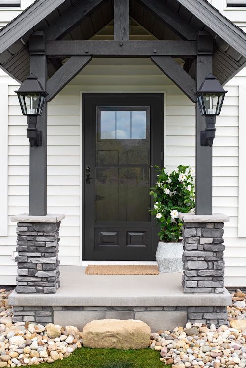 Front door with dark trim, stacked stone columns, white siding, and lantern lights.