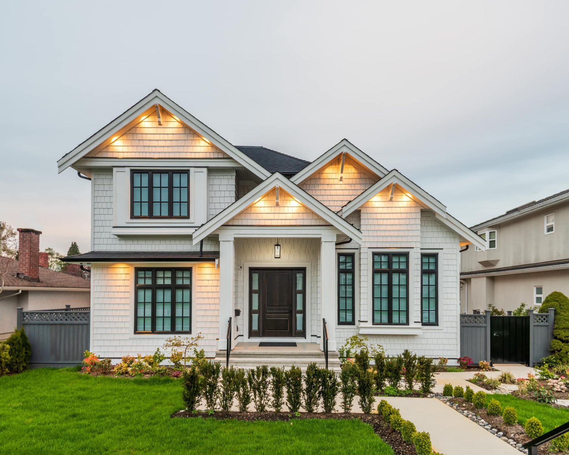 White two-story house with black-framed windows, lighted gables, and a well-manicured lawn and walkway.