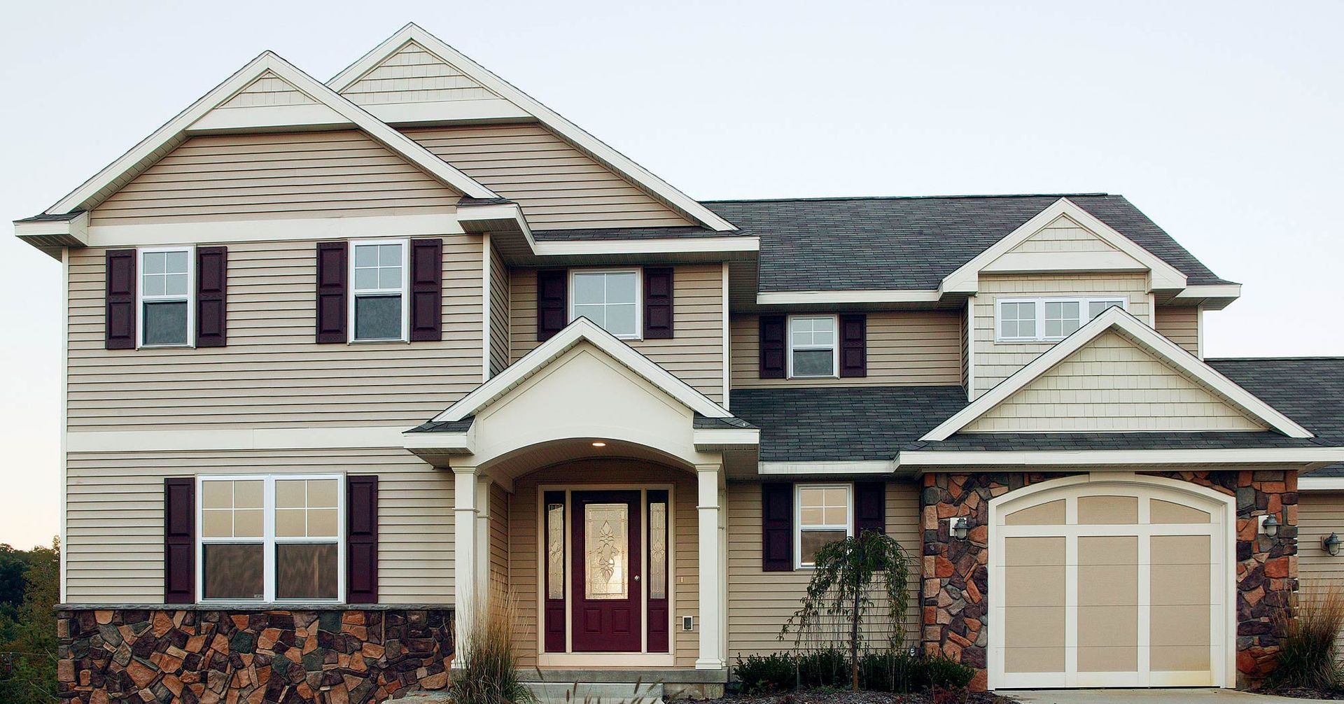 Two-story house with tan siding, burgundy shutters, and stone accents; a covered entryway with a burgundy door and a tan garage door.