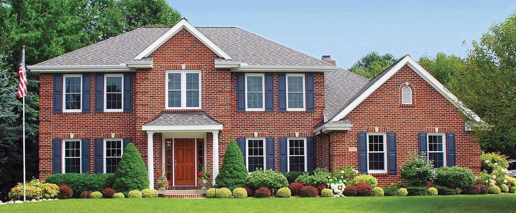 Red brick two-story house with blue shutters, a green lawn, and shrubbery. A US flag flies.