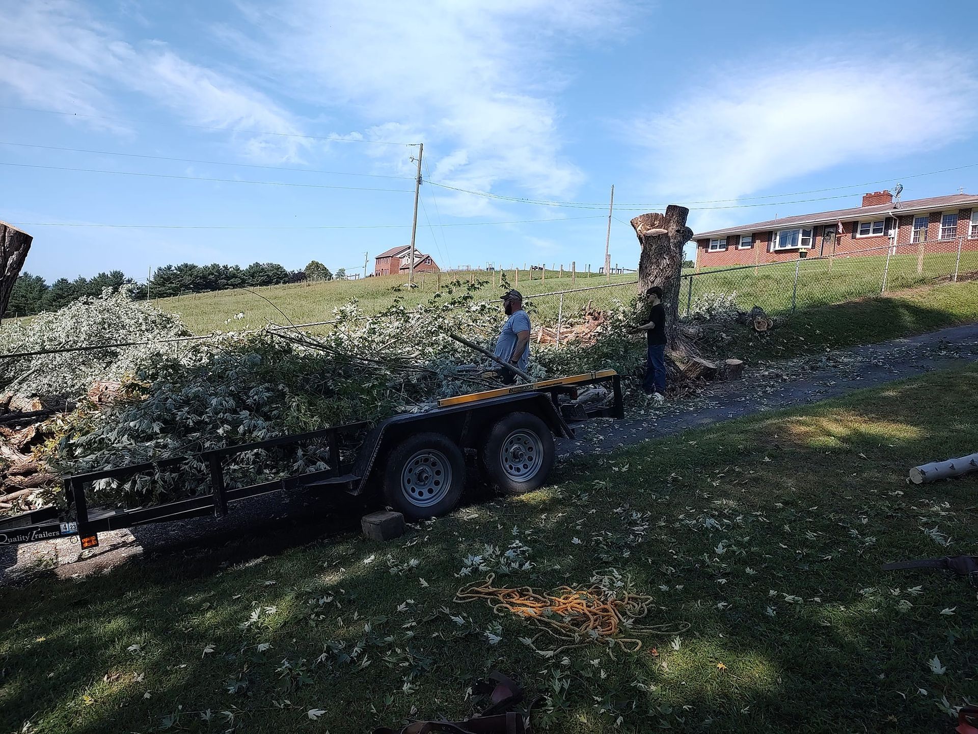 A person loads tree trimmings onto a flatbed trailer on a grassy slope near a brick house.