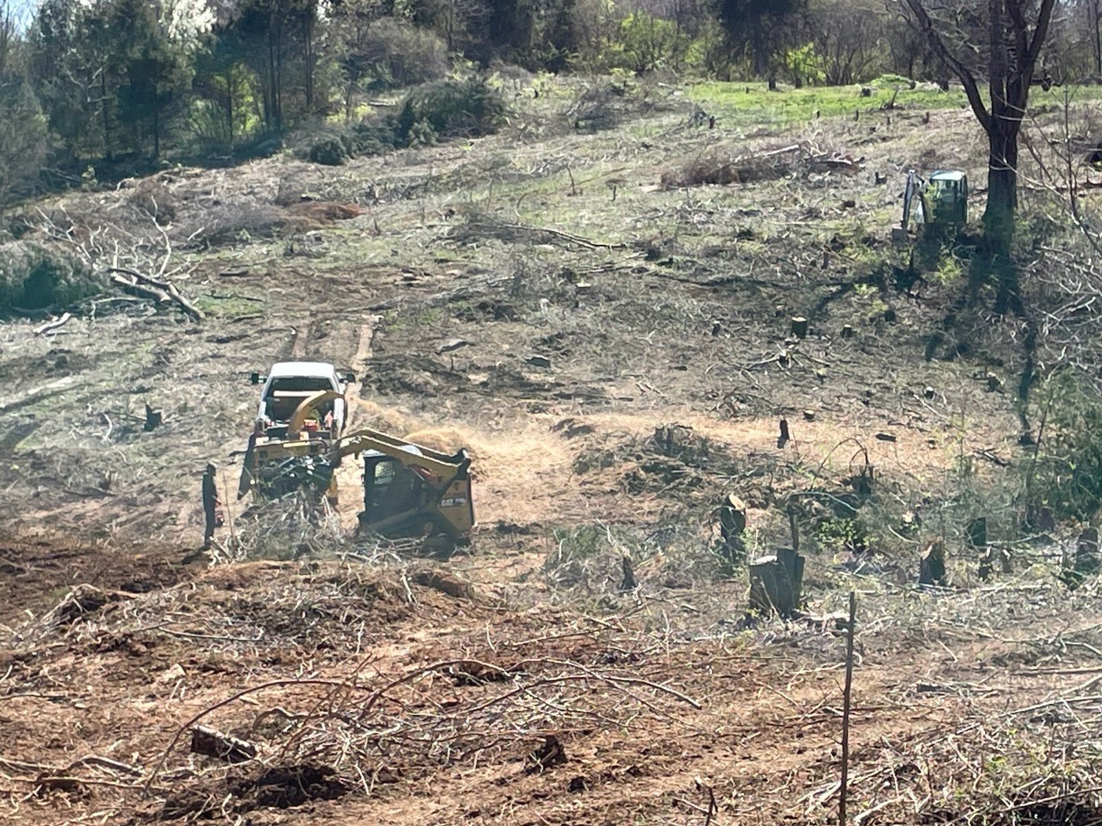 A yellow excavator and a white pickup truck work on a cleared, hilly landscape filled with tree stumps and brush.
