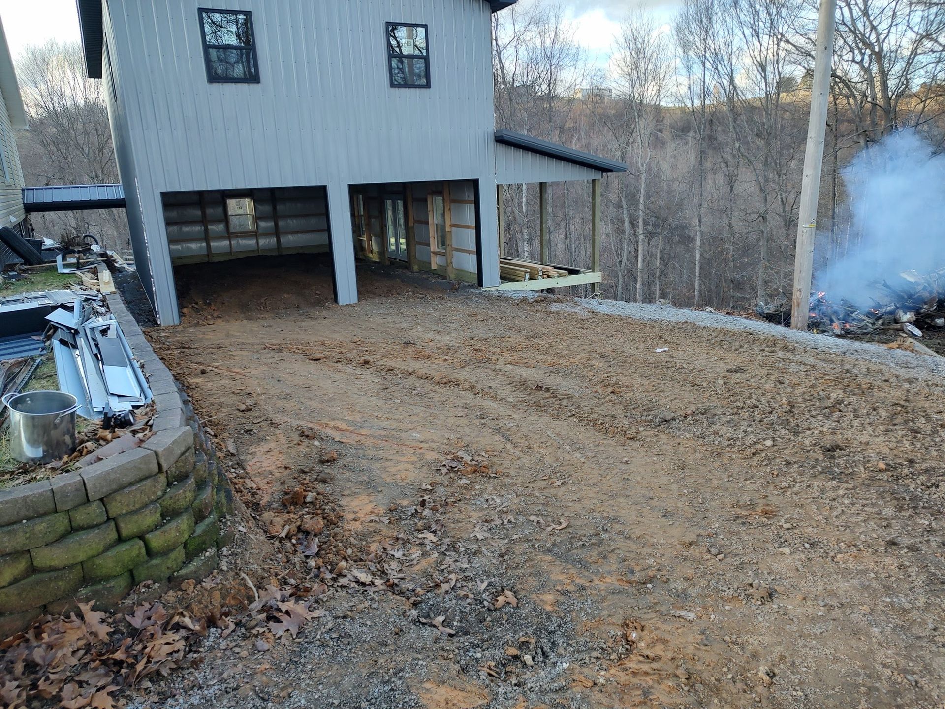 A gray house with an open ground level sits next to a dirt driveway, a stone retaining wall, and a plume of white smoke.