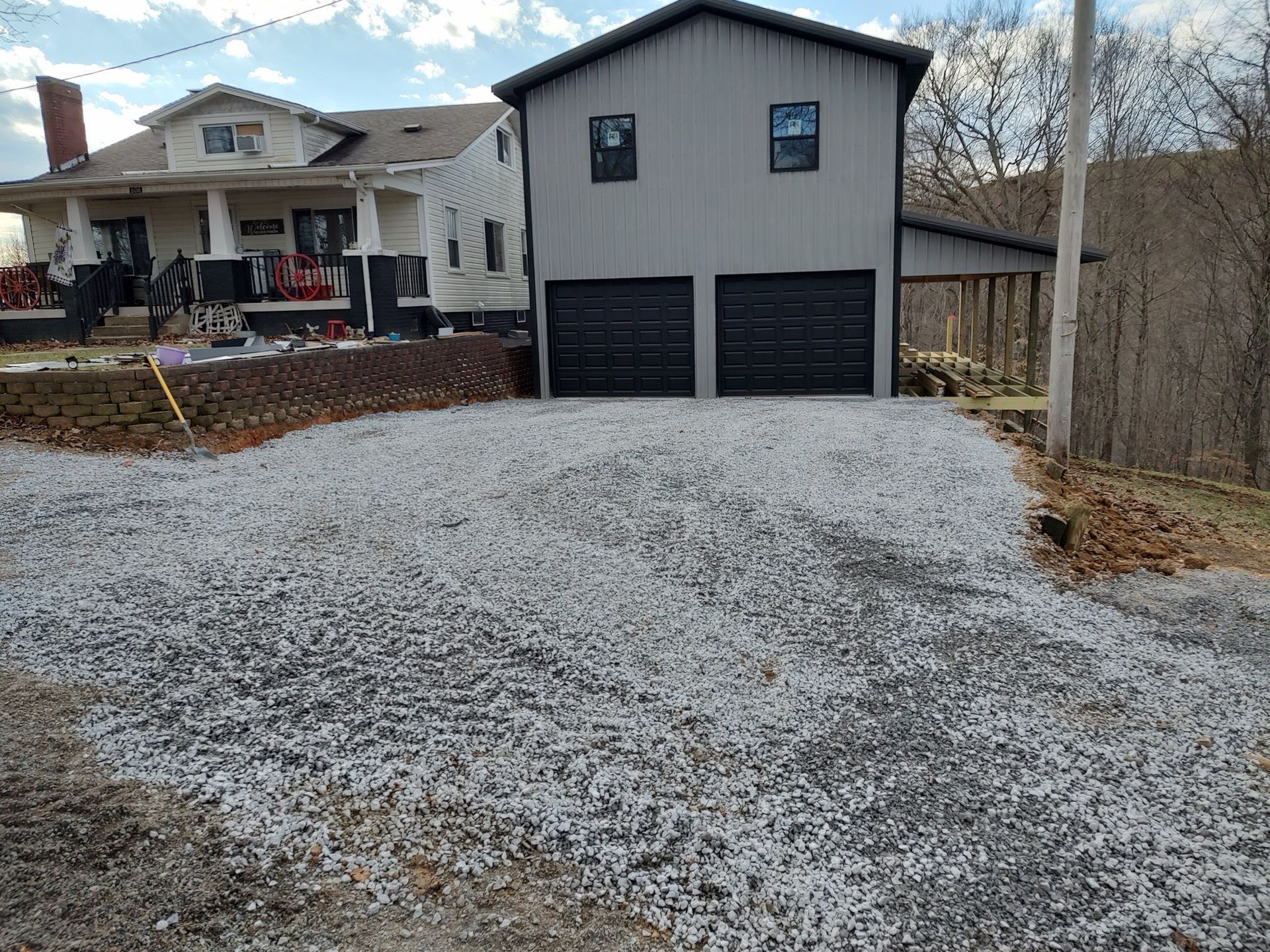 A gray two-story garage with black doors and a house with a porch stand behind a gravel driveway under a bright sky.