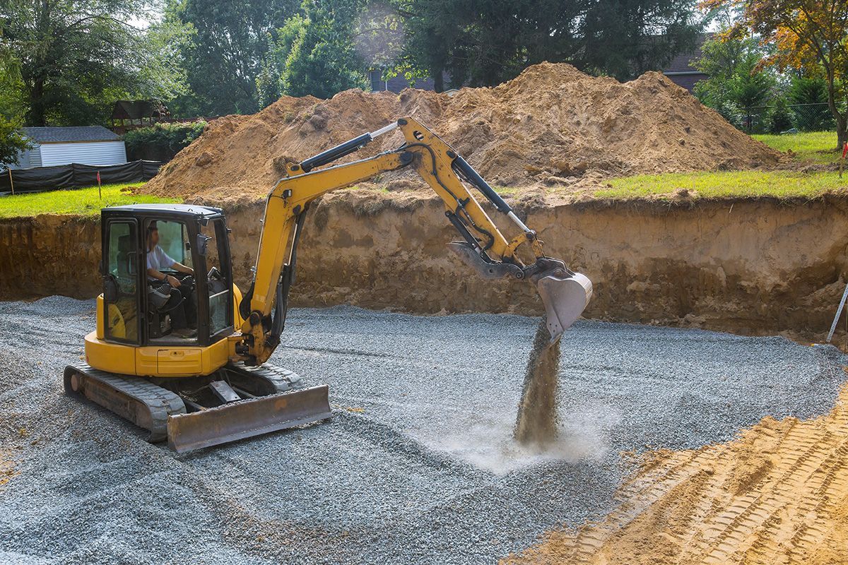 A small yellow excavator spreads gravel on the base of a deep, excavated construction pit outdoors.