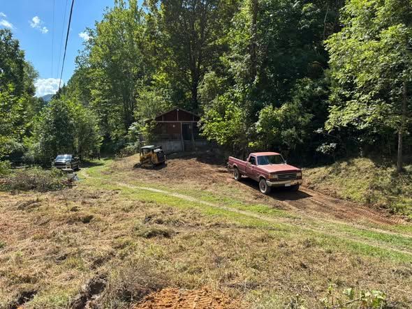 A reddish-brown truck and a small yellow excavator parked in a clearing near a rustic wooden shed in a wooded area.