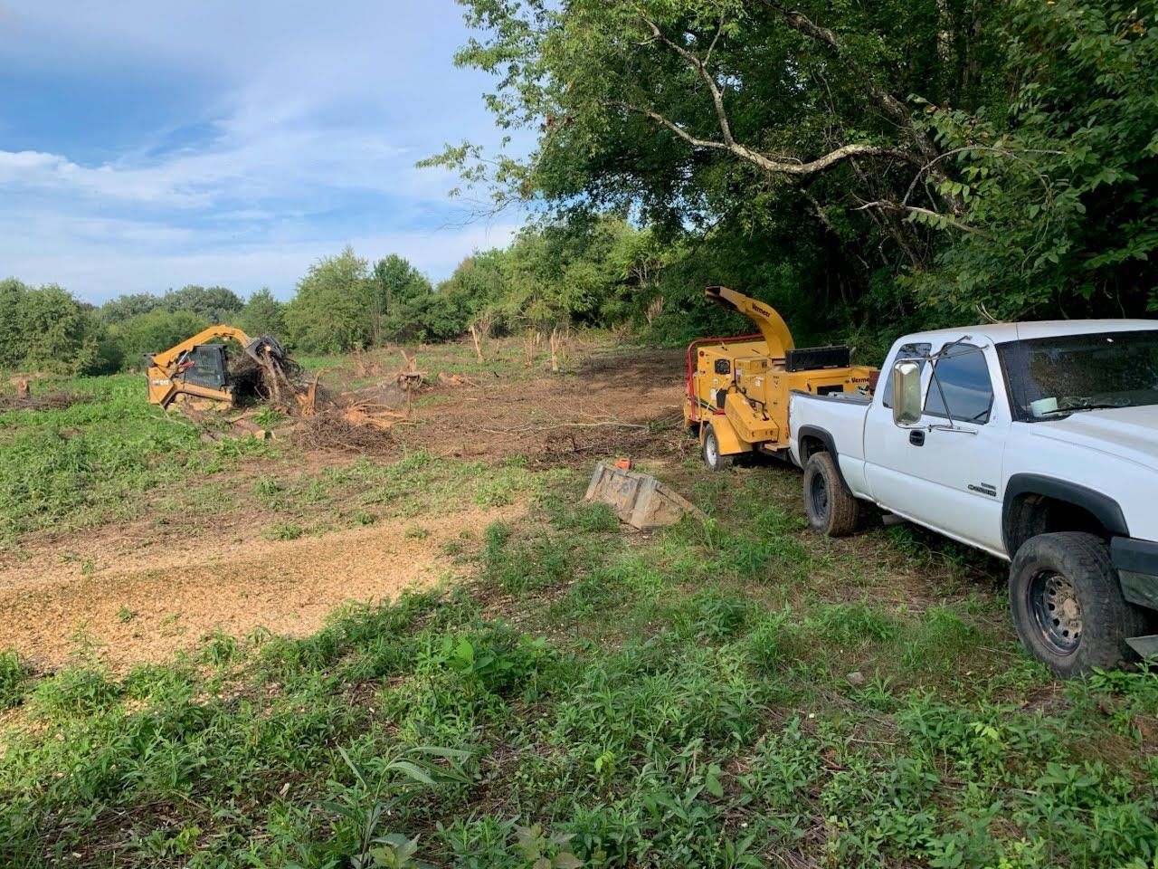 A white pickup truck pulling a yellow wood chipper, with a yellow skid steer in a cleared, grassy field.