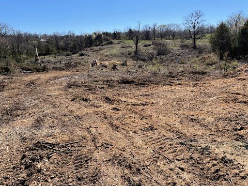 A cleared, rugged field of brown dirt with tire tracks and debris, surrounded by a line of trees under a clear blue sky.