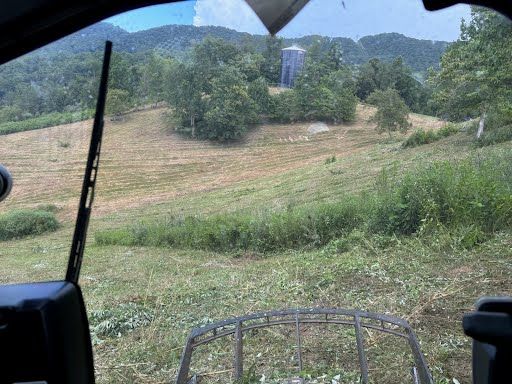 View from a vehicle windshield looking out over a grassy hillside with a distant structure and trees under a cloudy sky.