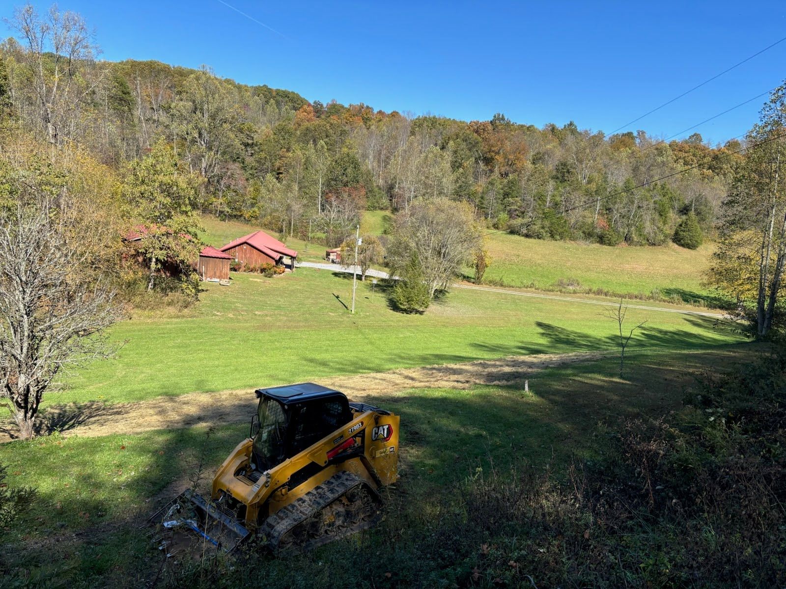 A yellow skid steer loader sits on a grassy hill overlooking a meadow with small buildings near a tree-lined mountain.