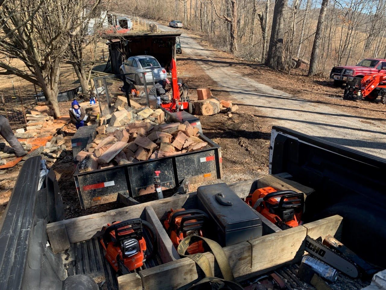 A truck bed holds multiple chainsaws, with a trailer of firewood, a log splitter, and wooded surroundings in the distance.